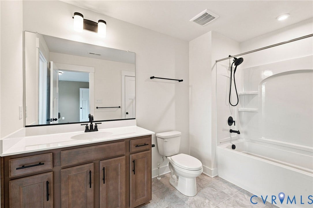 En-suite bathroom. Spacious bathroom featuring a single vanity with wood-finish cabinetry, a large frameless mirror, a white bathtub with shower, and tiled flooring