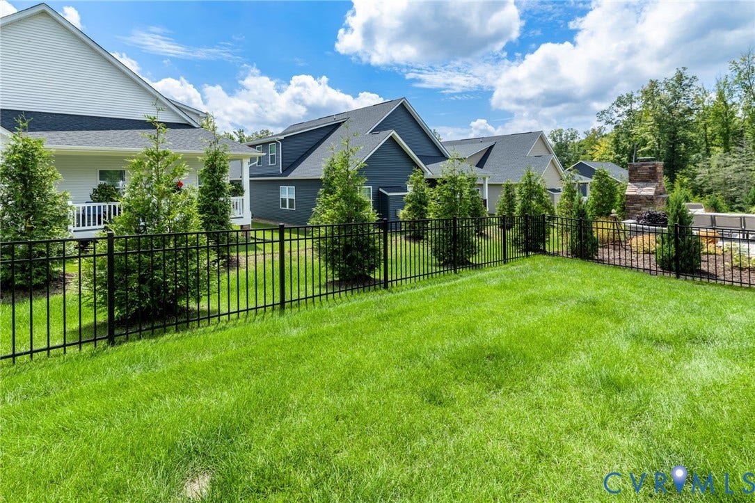 6249 Deer Summit Court, Charleston Club, Moseley, VA 23120 Expansive green lawn with a black metal fence, mature plantings, and a stone outdoor fireplace