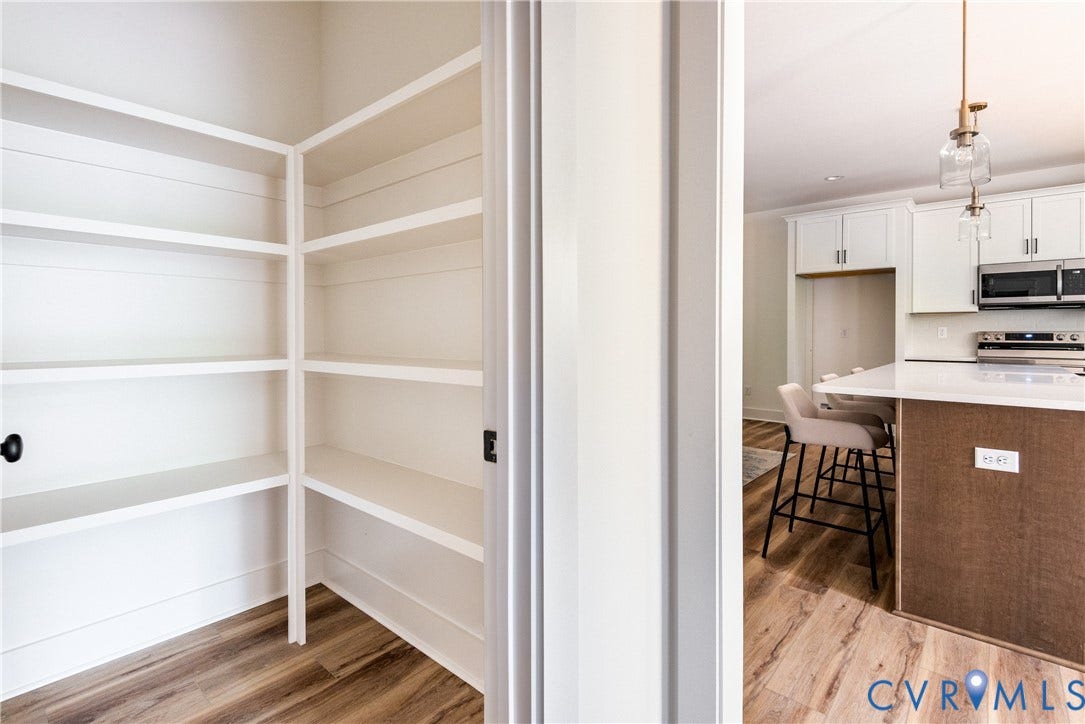 Walk-in pantry with extensive shelving, LVP flooring, and a view into the kitchen featuring white cabinetry, an island with a light-toned countertop, and stainless steel appliances
