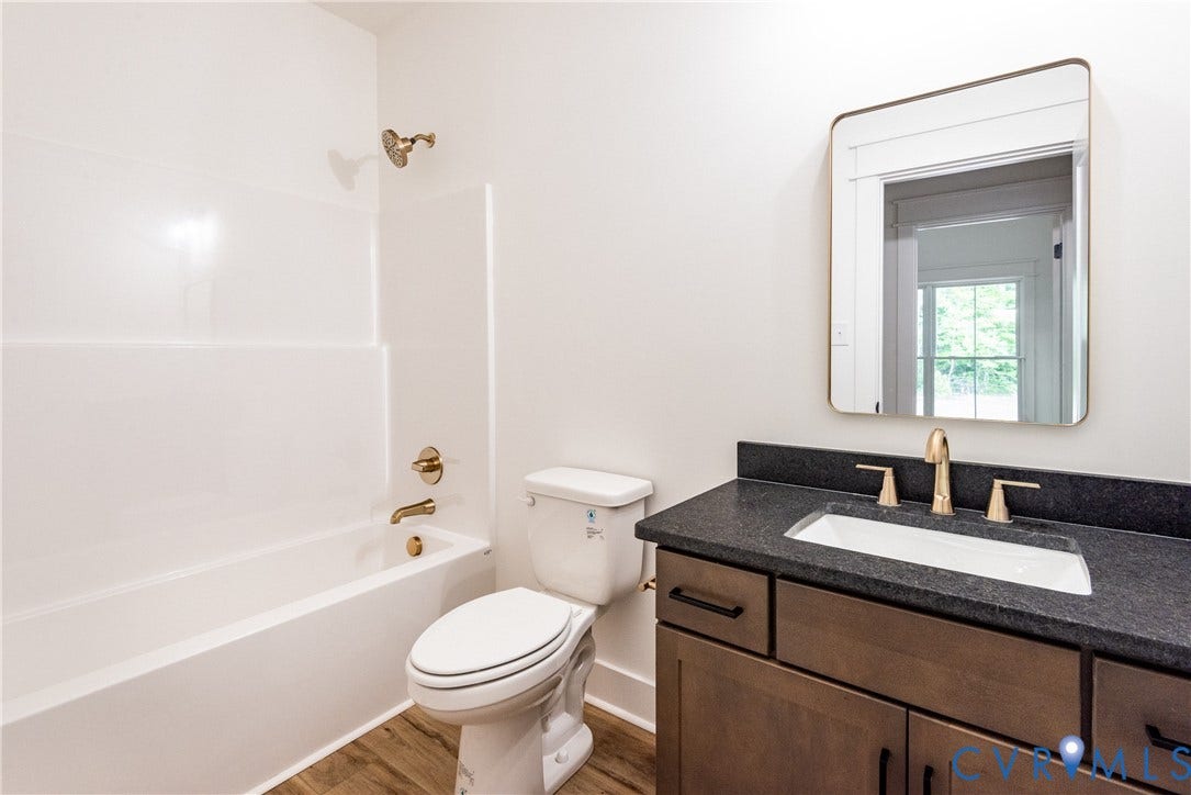 Bathroom featuring a white tub with integrated shower, vanity with a dark countertop and undermount sink, brass finish fixtures, LVP flooring, and a framed wall mirror