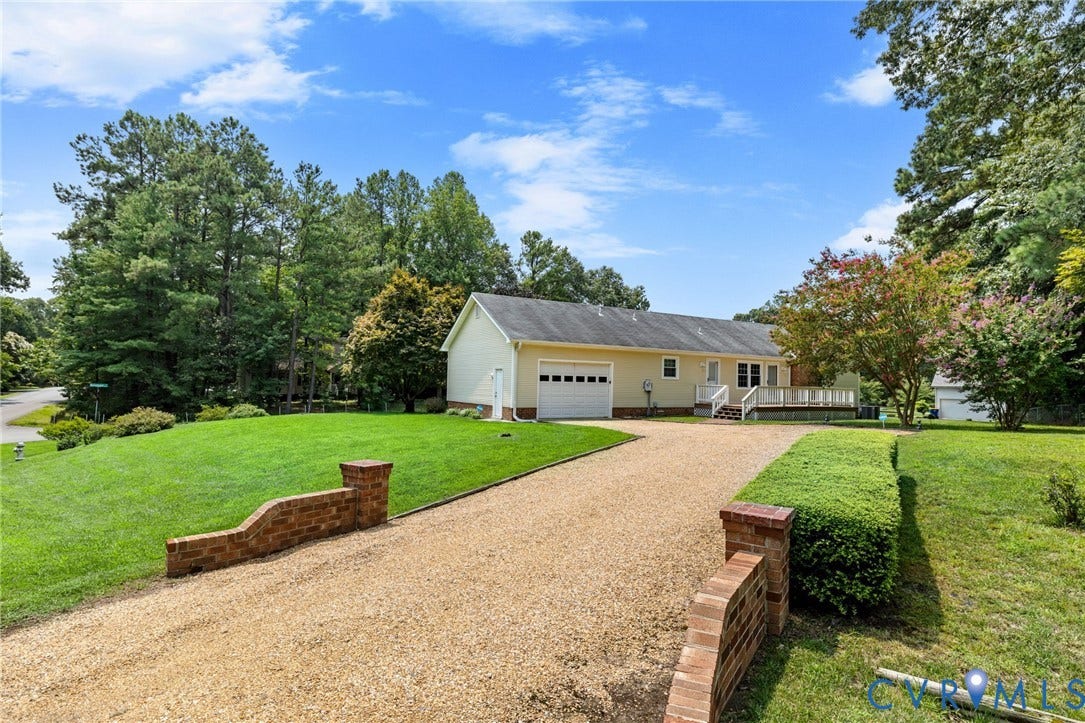 Ranch-style residence featuring a gravel driveway, brick entry walls, an attached garage, a spacious rear deck, and mature landscaping