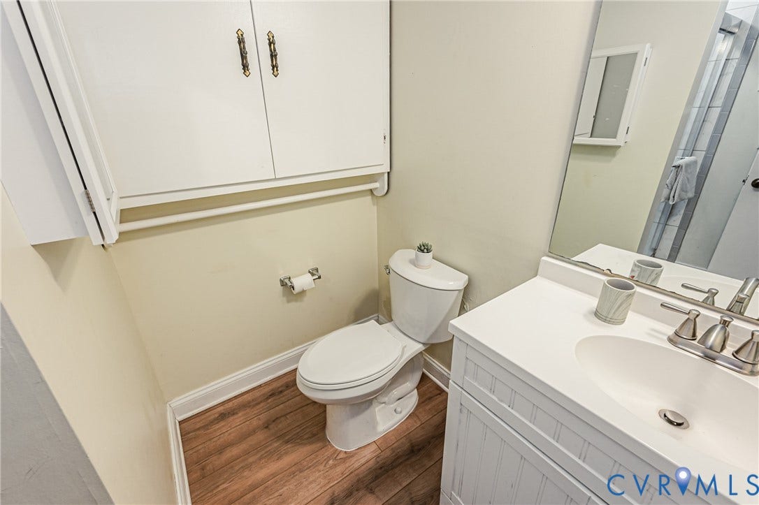 9406 Emmett Court, Glen Allen, VA 23060 Bathroom featuring a white porcelain toilet with tank, pedestal sink with chrome faucet and mirror, wood-finish flooring, white cabinetry with brushed nickel hardware, and neutral wall finishes throughout