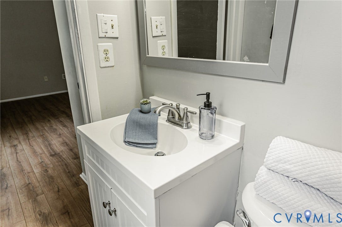 9406 Emmett Court, Glen Allen, VA 23060 Clean and functional bathroom vanity featuring a white pedestal sink with chrome faucet and fixtures, ample countertop space, white cabinetry with hardware, a framed mirror, and wood-finish flooring throughout the space