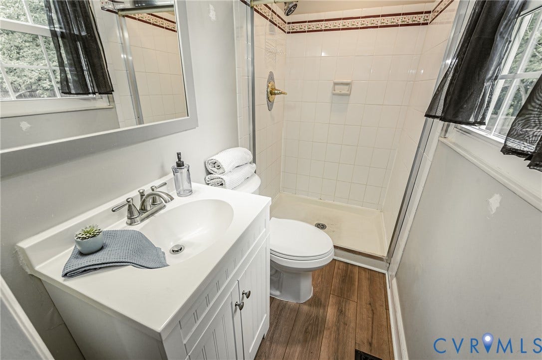 9406 Emmett Court, Glen Allen, VA 23060 Bathroom featuring a white vanity with integrated sink, brushed nickel faucet, wood-finish flooring, a white tiled shower with glass enclosure, and a window providing natural light