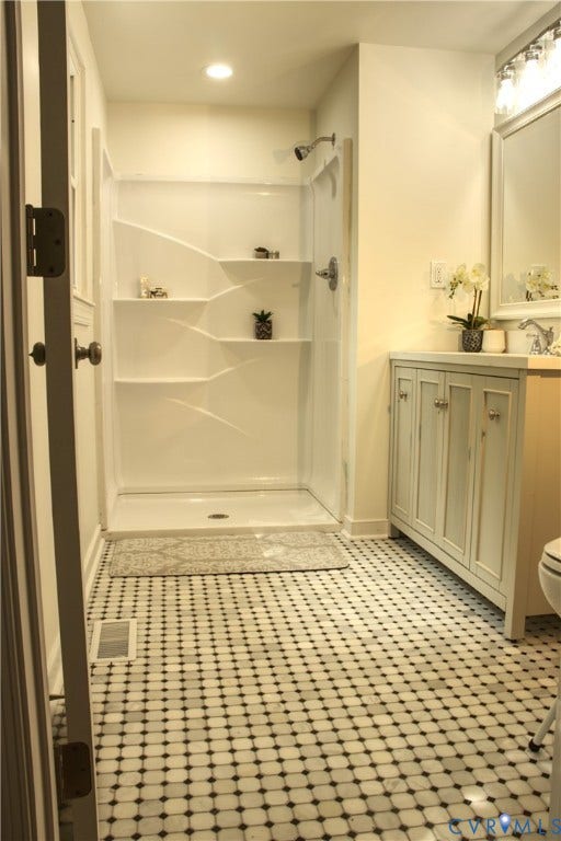 5004 Fairlake Lane, Henrico, VA 23060 White tiled shower stall with built-in shelving, black and white mosaic floor tiling, light-colored vanity with built-in cabinetry, and wall-mounted lighting fixture