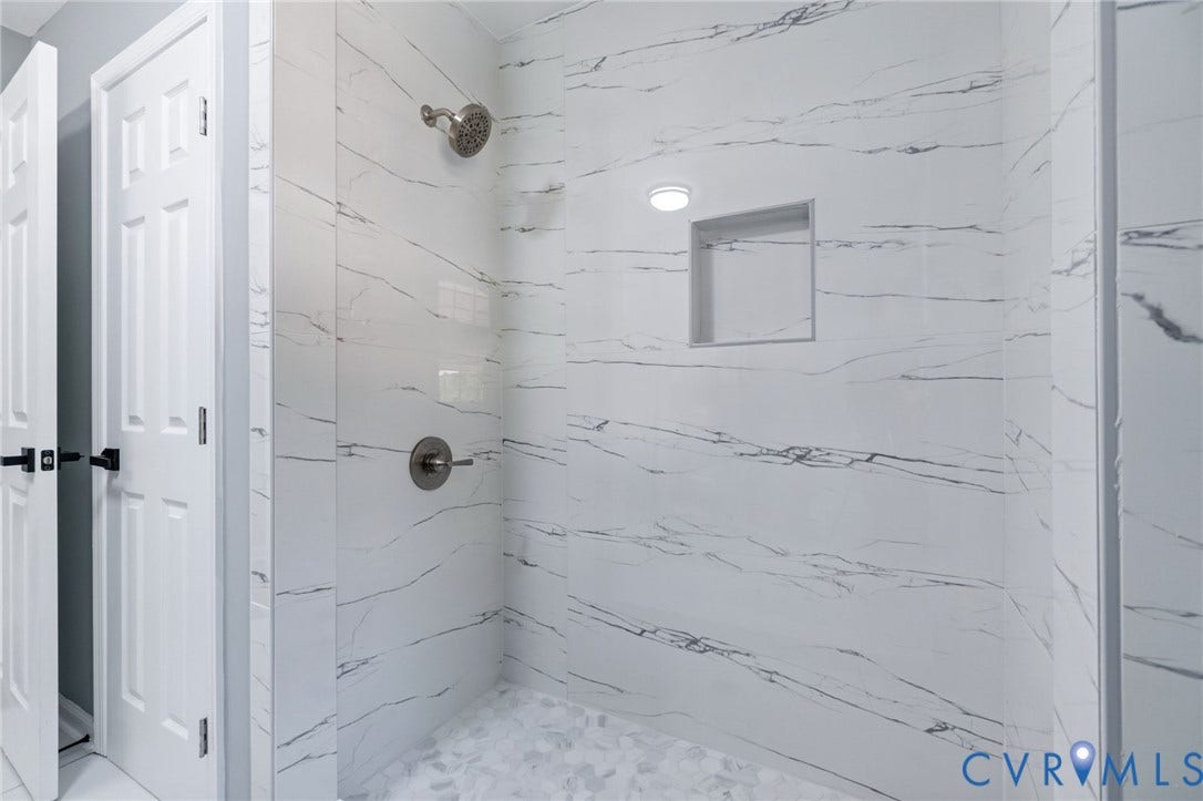 Shower stall featuring white marble-patterned wall tile, a recessed shower niche, brushed nickel shower fixtures, and white pebble-style floor tile