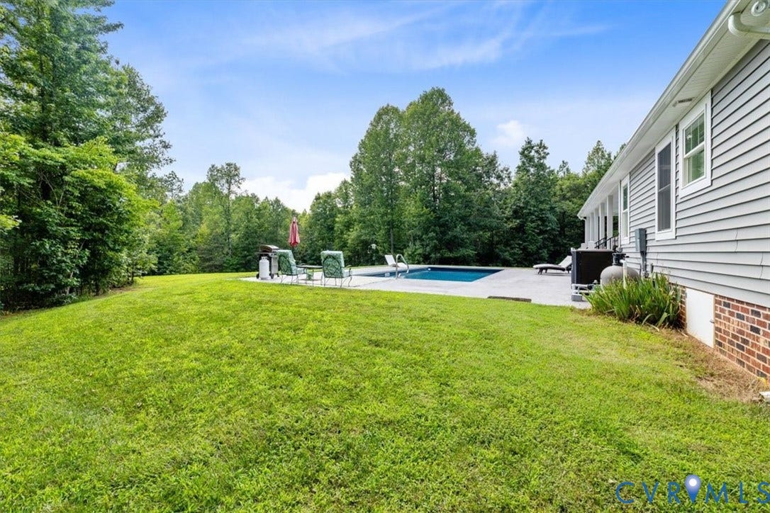 Expansive lawn area featuring a rectangular pool with a surrounding paved deck, bordered by mature trees