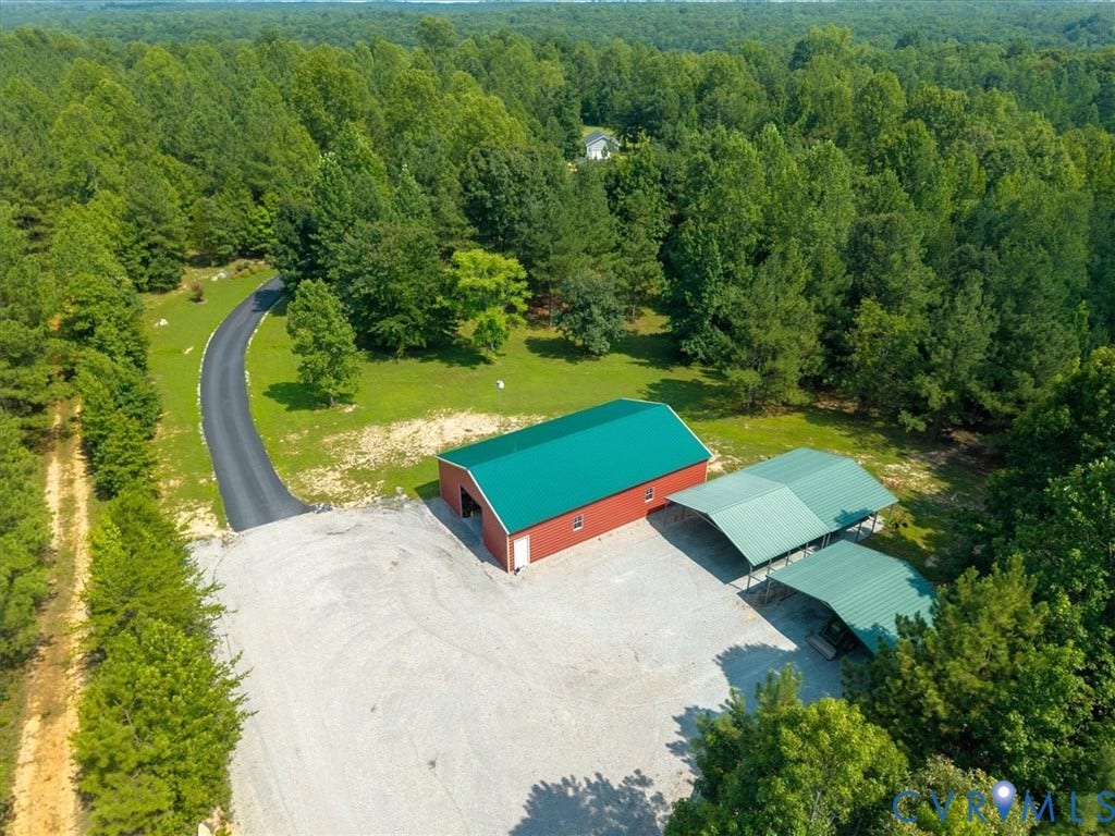 Red outbuilding with a green metal roof, gravel lot, two green metal carports, and a paved driveway surrounded by mature trees