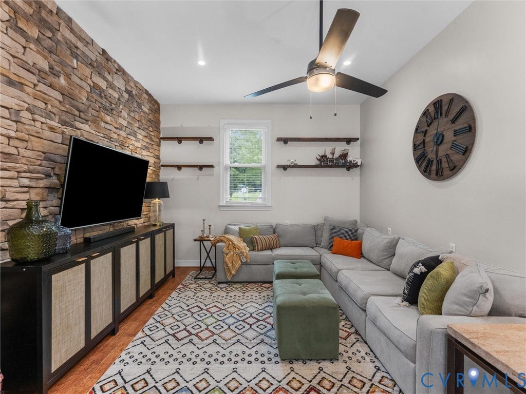 8216 Indian Springs Road, Richmond, VA 23237 Bright, open living room featuring exposed brick accent wall with mounted flat-screen television, ceiling fan with integrated lighting, hardwood flooring, geometric patterned area rug, gray upholstered sectional sofa with throw pillows, built-in shelving, and large window providing natural light