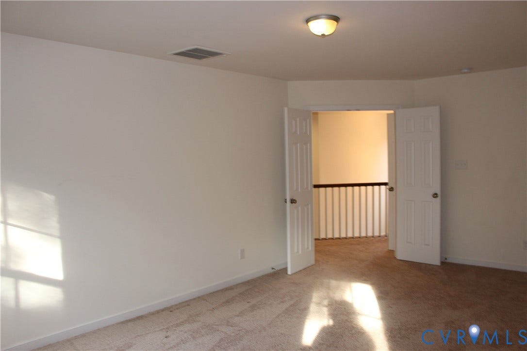 Carpeted room with white walls, a flush-mount ceiling light, and white double doors opening to a hallway with a white railing