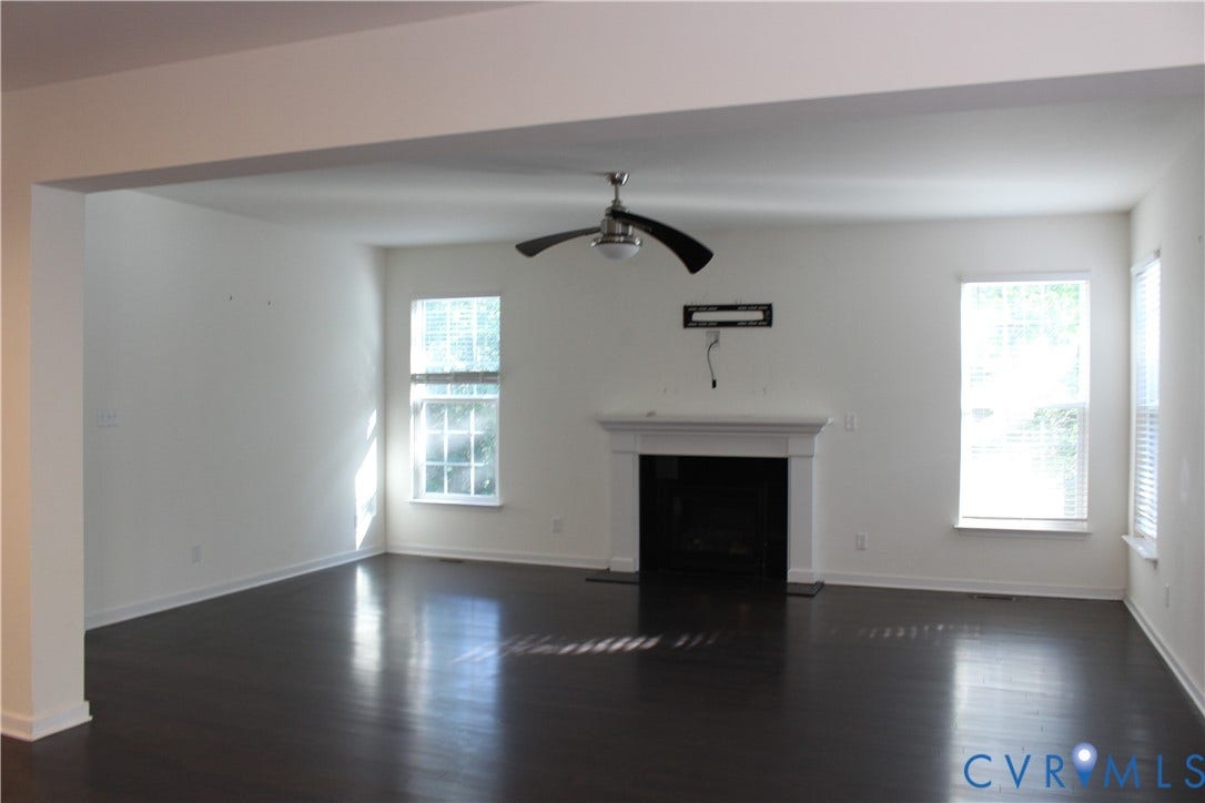 Spacious living area featuring dark wood-finish flooring, a classic white mantel fireplace, multiple windows with blinds, and a ceiling fan with light fixture