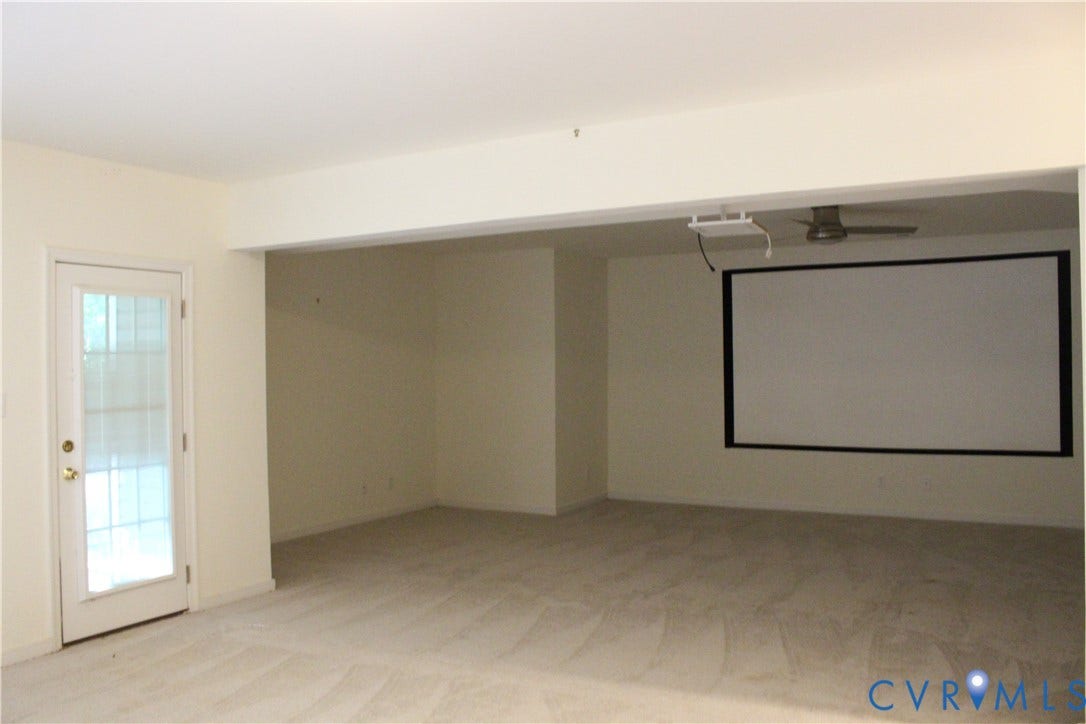 Spacious room featuring a ceiling-mounted projector and large screen, light-colored carpeting, recessed lighting, and a glass-paneled door