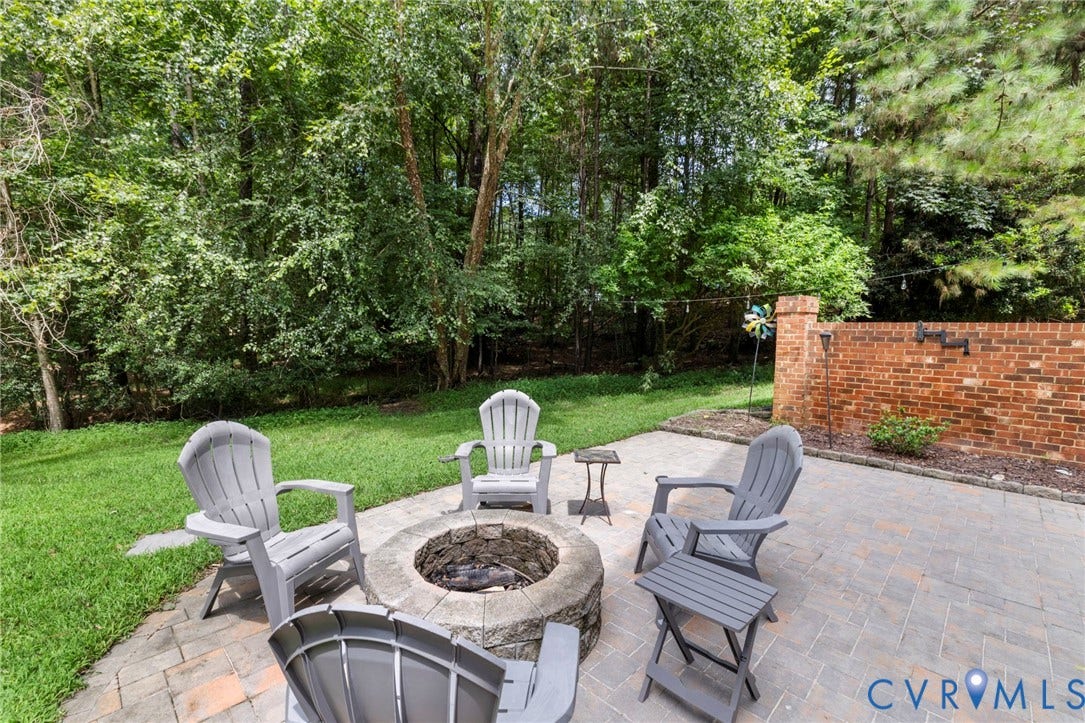 8343 Shagreen Court, The Highlands, Chesterfield, VA 23838 Paver patio with a stone fire pit, surrounded by lush green lawn and mature trees, featuring a partial brick wall