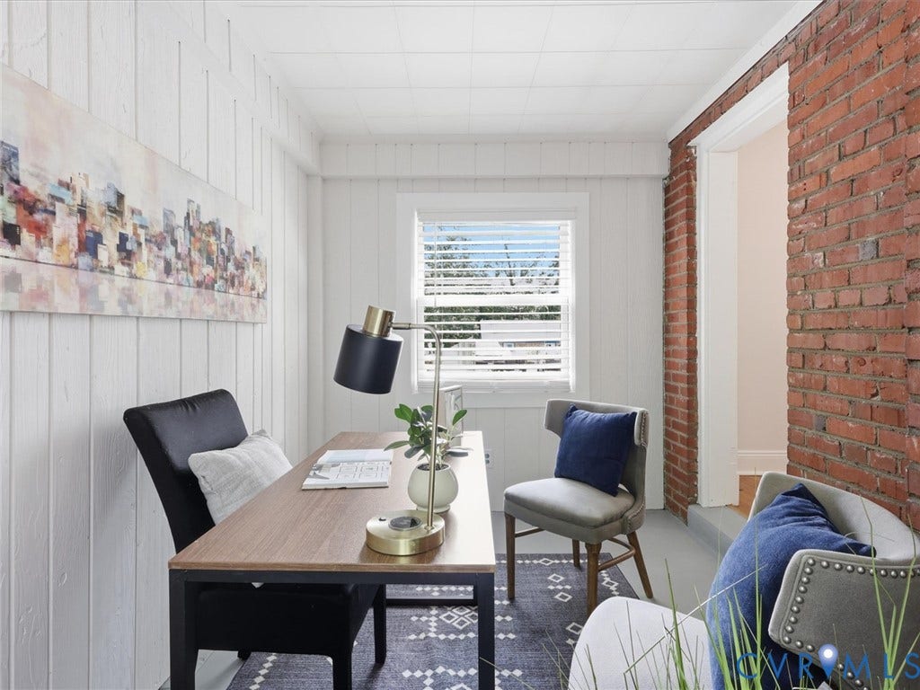 Vertical paneling, exposed brick accent wall, window with blinds, wood-finish desk, and a textured area rug