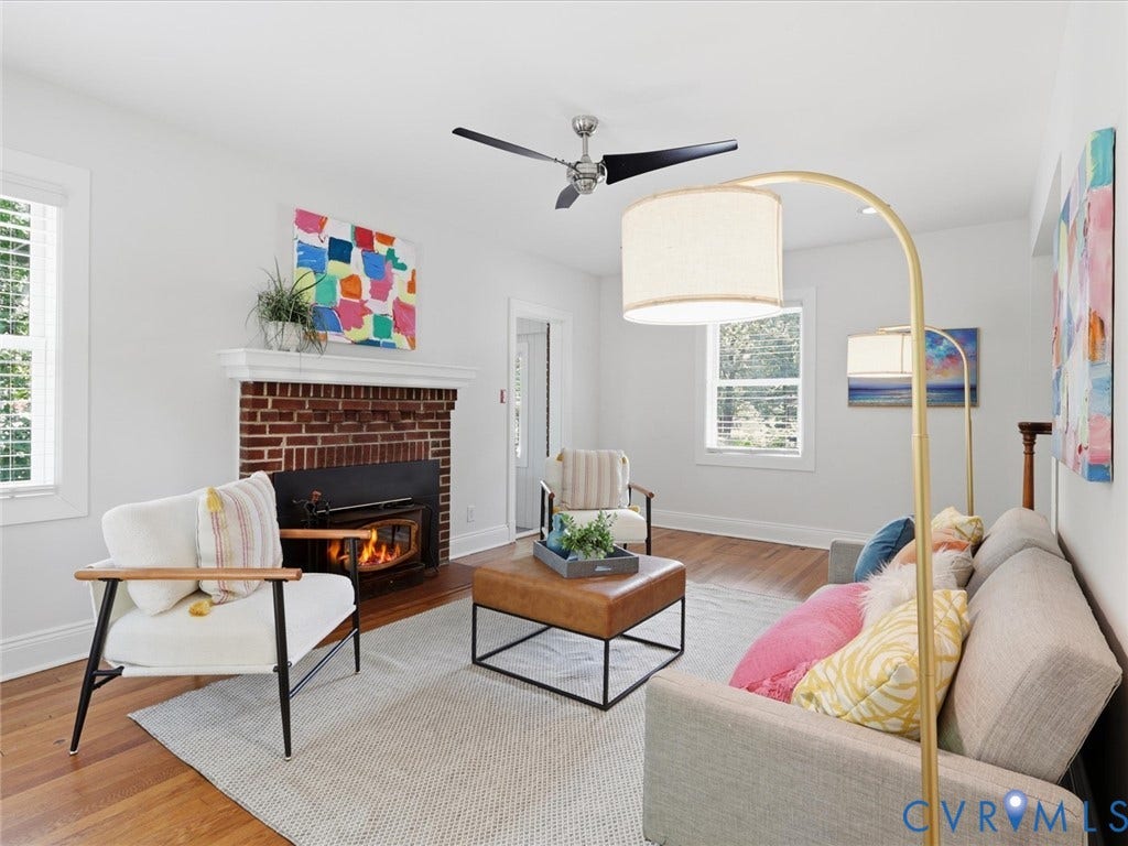 Brick hearth fireplace with white mantel, hardwood flooring, ceiling fan, and multiple windows with blinds