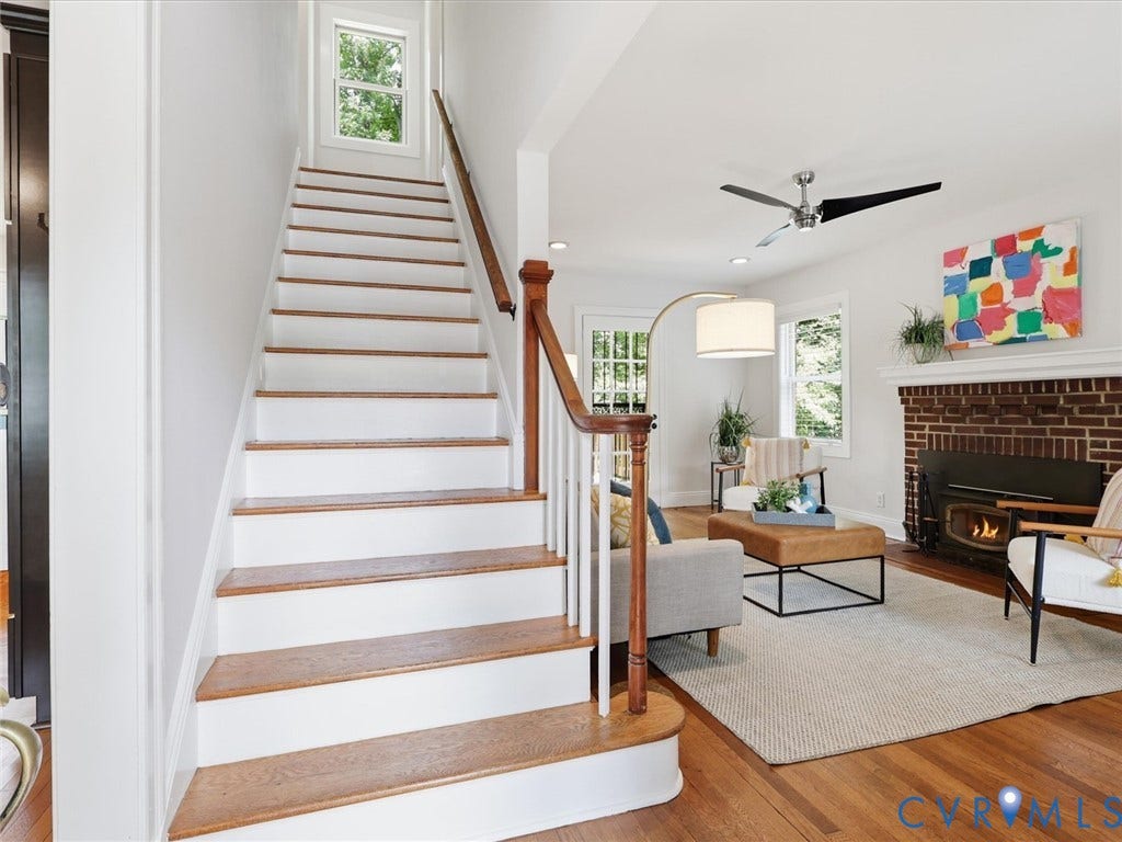 Hardwood flooring, brick fireplace with insert, white painted wood mantel, modern ceiling fan, and staircase with wood treads and banister