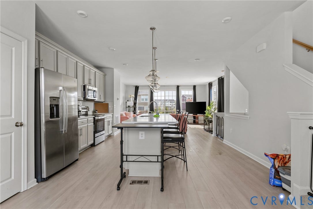 Open-concept kitchen and dining area featuring stainless steel appliances, white cabinetry with hardware accents, a large center island with seating, wood-finish flooring throughout, pendant lighting, and abundant natural light from multiple windows