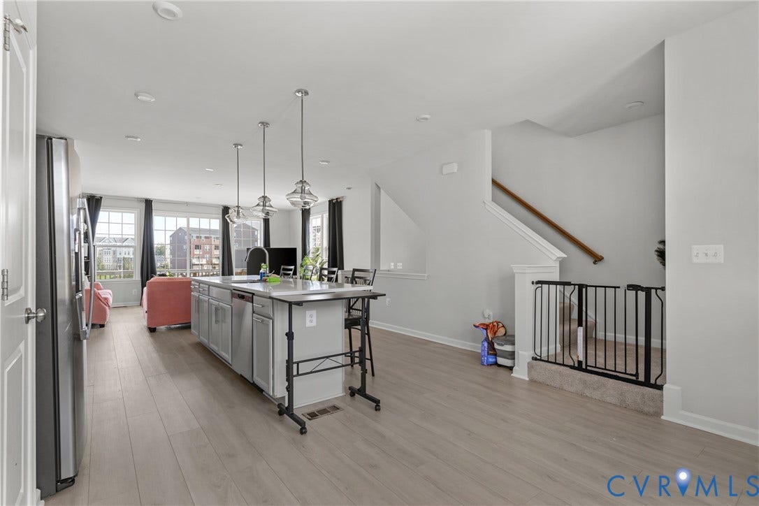 Open-concept kitchen and living area featuring stainless steel appliances, gray wood-finish flooring throughout, white shaker-style cabinetry with island prep space, pendant lighting, and abundant natural light from multiple windows