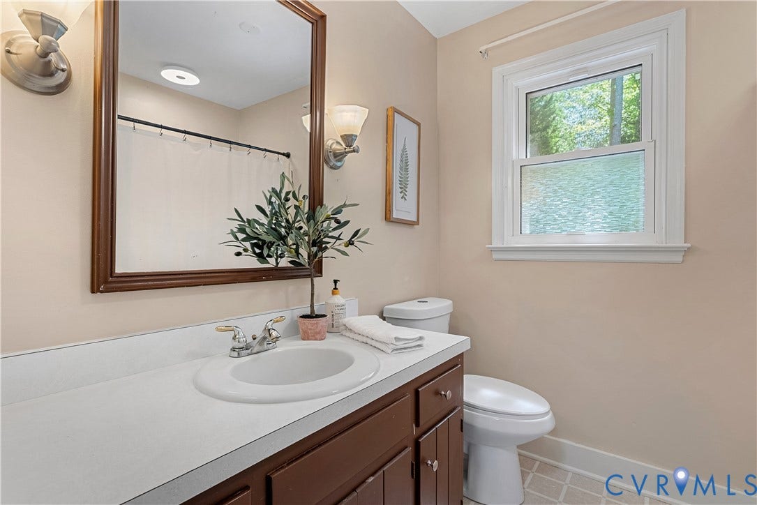 Elegant bathroom featuring a curved pedestal sink with chrome faucet, wood-finish vanity cabinetry with ample storage, white subway tile wainscoting, framed mirror with bronze-finish trim, recessed ceiling lighting, and a casement window providing natural light