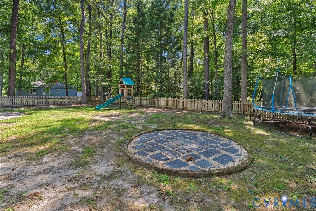Expansive backyard oasis nestled among mature hardwood trees, featuring a circular stone fire pit, manicured lawn, wooden perimeter fencing, and a multi-level play structure with slide