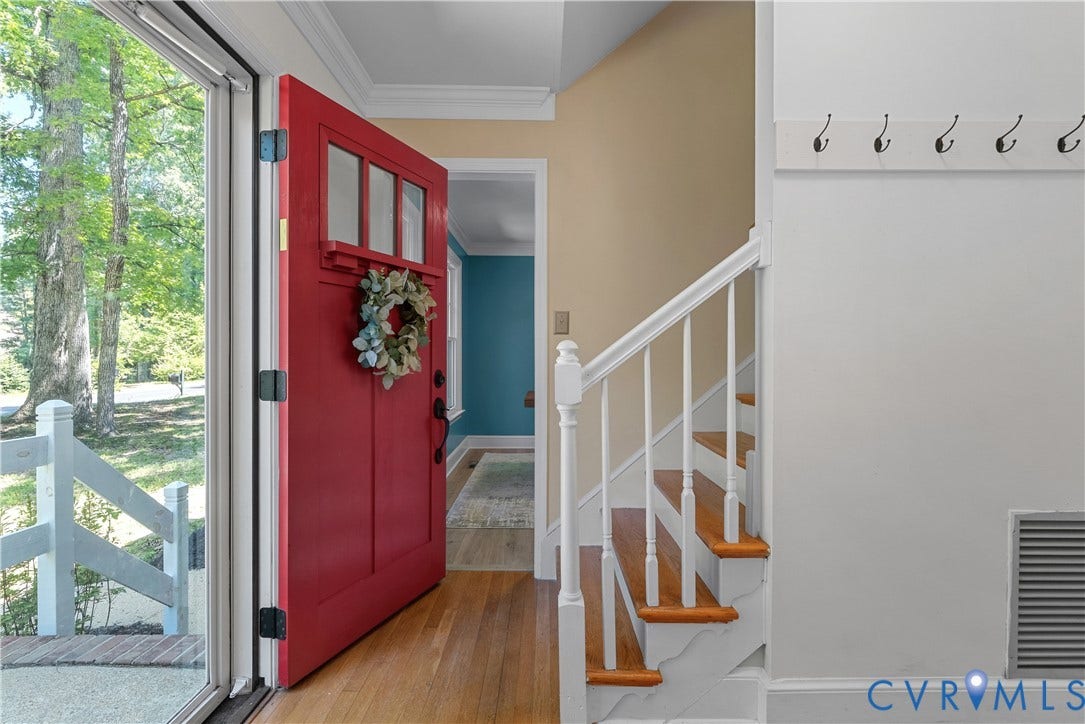 Welcoming foyer featuring a striking red entry door with transom window and festive wreath, warm wood-finish flooring, white painted staircase with wood-tone treads and metal railings, and abundant natural light from expansive glass doors overlooking mature landscaping