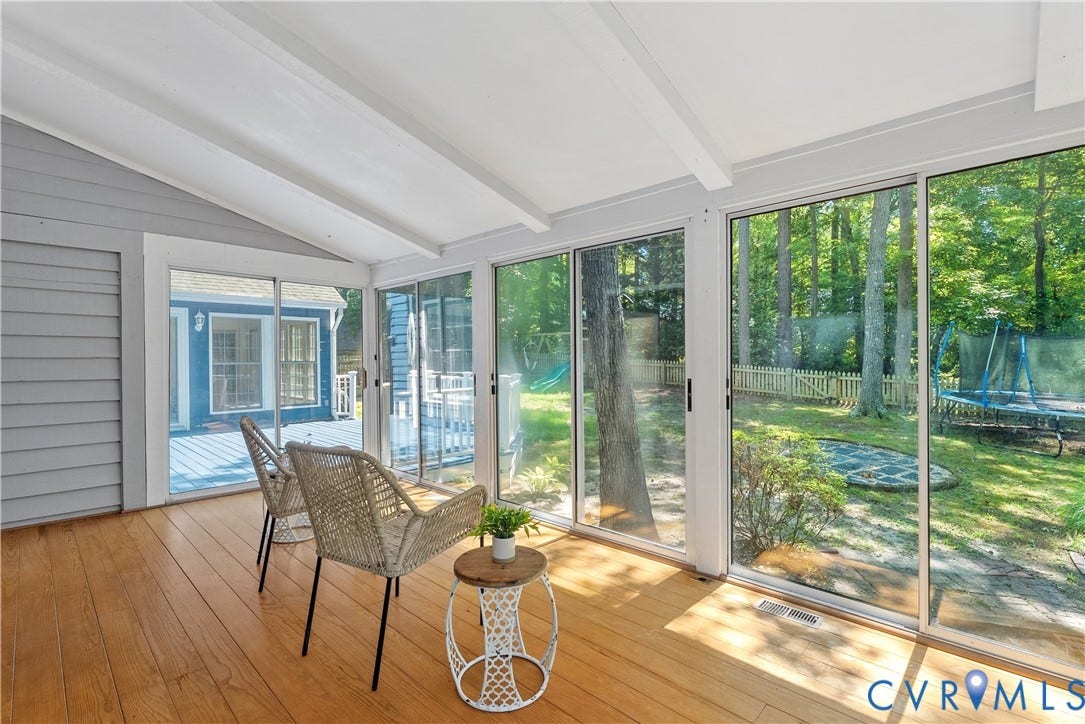 Bright sunroom with expansive glass sliding doors and windows framing lush outdoor scenery, warm wood-finish flooring, white coffered ceiling with exposed beams, and direct access to a waterfront setting with mature landscaping and dock infrastructure