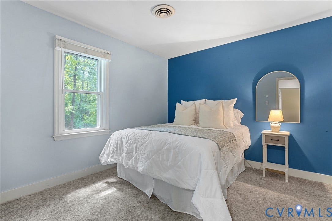 Bright bedroom featuring a large double-hung window with white trim, soft blue accent wall, neutral carpet flooring, and a coordinating bedside table with integrated lighting fixture