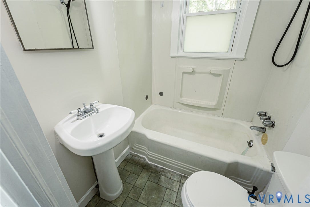 Pedestal sink with chrome faucet, bathtub with integrated shower, window with privacy screen, white wall paneling, and patterned floor tiles
