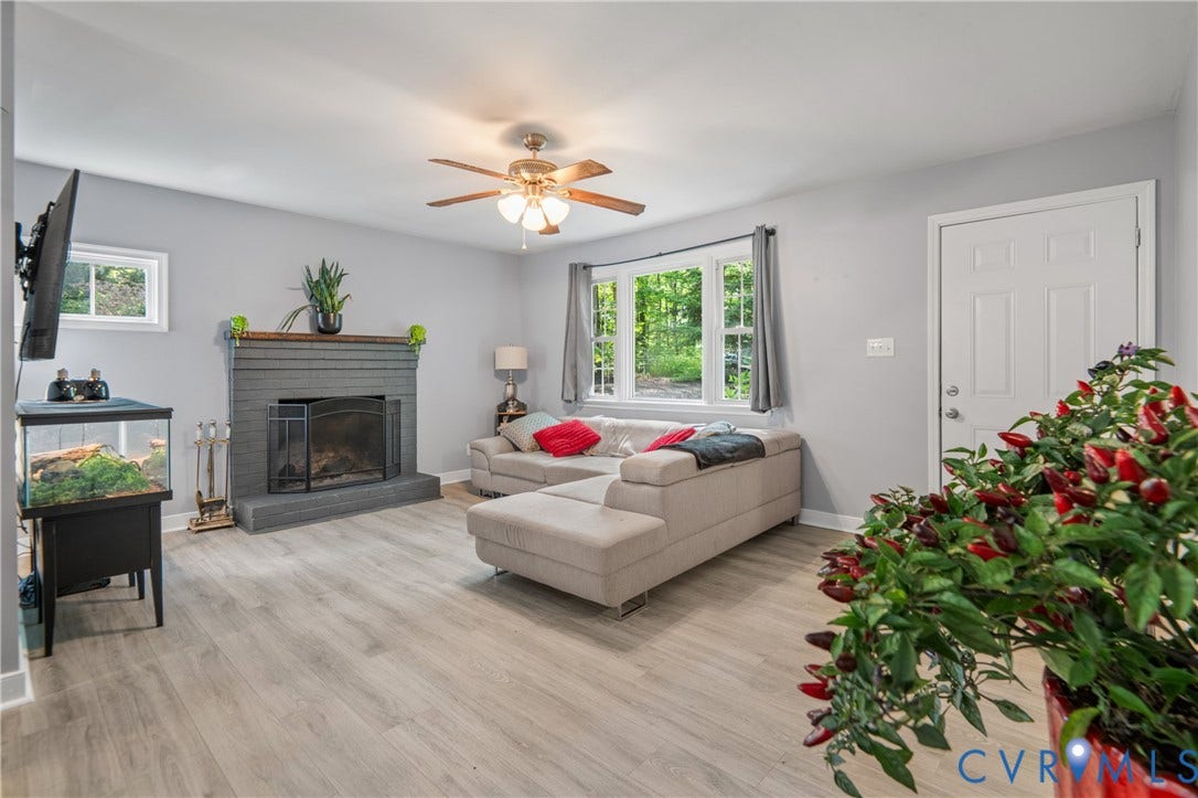 3940 Three Bridge Road, Powhatan, VA 23139 Gray-painted brick fireplace with a wood mantle, ceiling fan with integrated lighting, multiple windows, and wood-finish flooring
