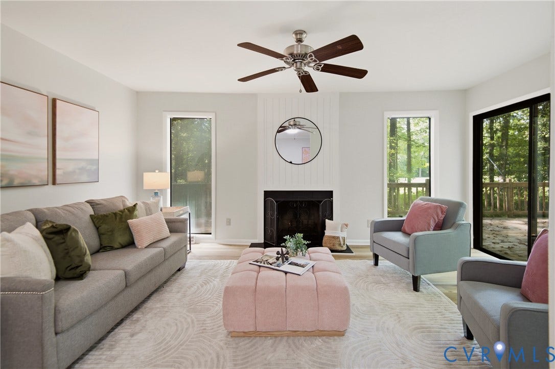 2903 Fox Chase Drive, Huntsbridge, Midlothian, VA 23112 Bright, light-filled living room featuring expansive glass doors and windows framing natural outdoor scenery, a wood-finish ceiling fan with brushed metal accents, a fireplace with black surround, and neutral-toned wood-finish flooring throughout