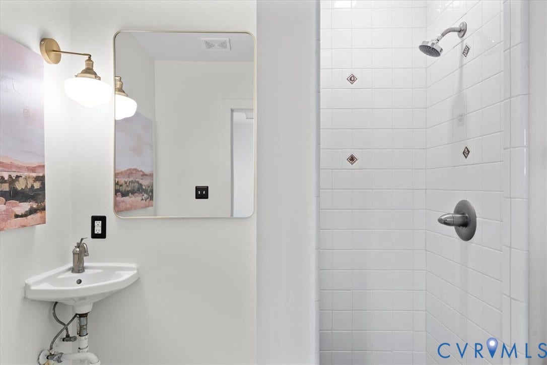 2510 E Leigh Street, Church Hill, Richmond, VA 23223 White tile shower with decorative accent tiles, corner pedestal sink, and rectangular framed mirror with wall-mounted brass light fixture
