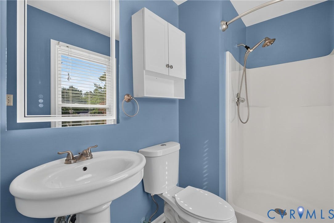 2020 Knotty Drive, Glen Allen, VA 23060 Bright, spa-like bathroom featuring soft blue walls, an oval pedestal sink with polished chrome fixtures, a white wall-mounted cabinet for storage, a walk-in shower with white tile surround and rainfall showerhead, and a low-flow toilet