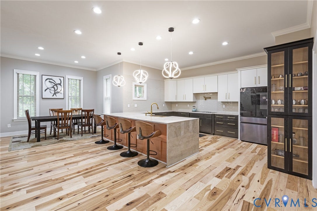 12554 Kain Road, Glen Allen, VA 23059 Open-concept kitchen and dining area featuring white cabinetry, a large waterfall-edge island with seating, stainless steel appliances, wood-finish flooring throughout, and abundant recessed and pendant lighting