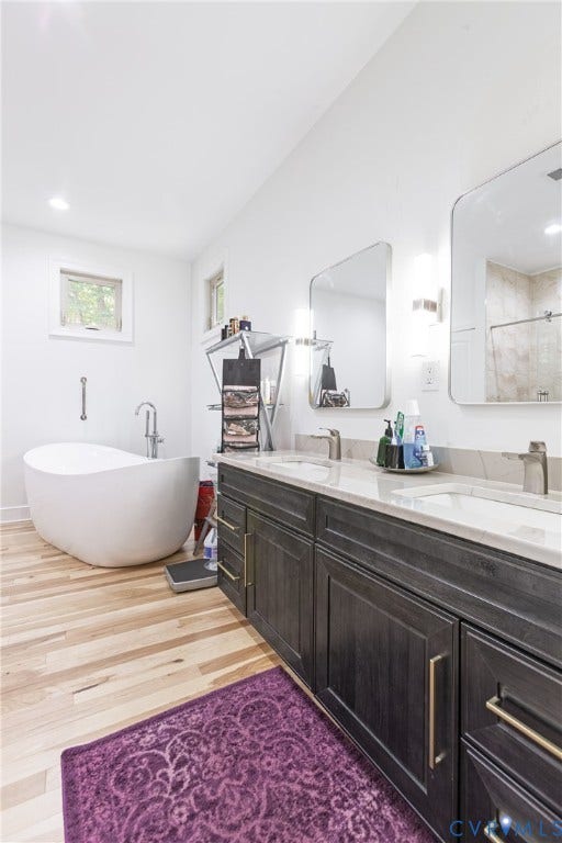 12554 Kain Road, Glen Allen, VA 23059 Spa-inspired primary bathroom featuring a freestanding soaking tub, dual vanity with dark cabinetry and brushed gold hardware, large framed mirrors with integrated lighting, wood-finish flooring, and a walk-in shower with glass enclosure and tile surround