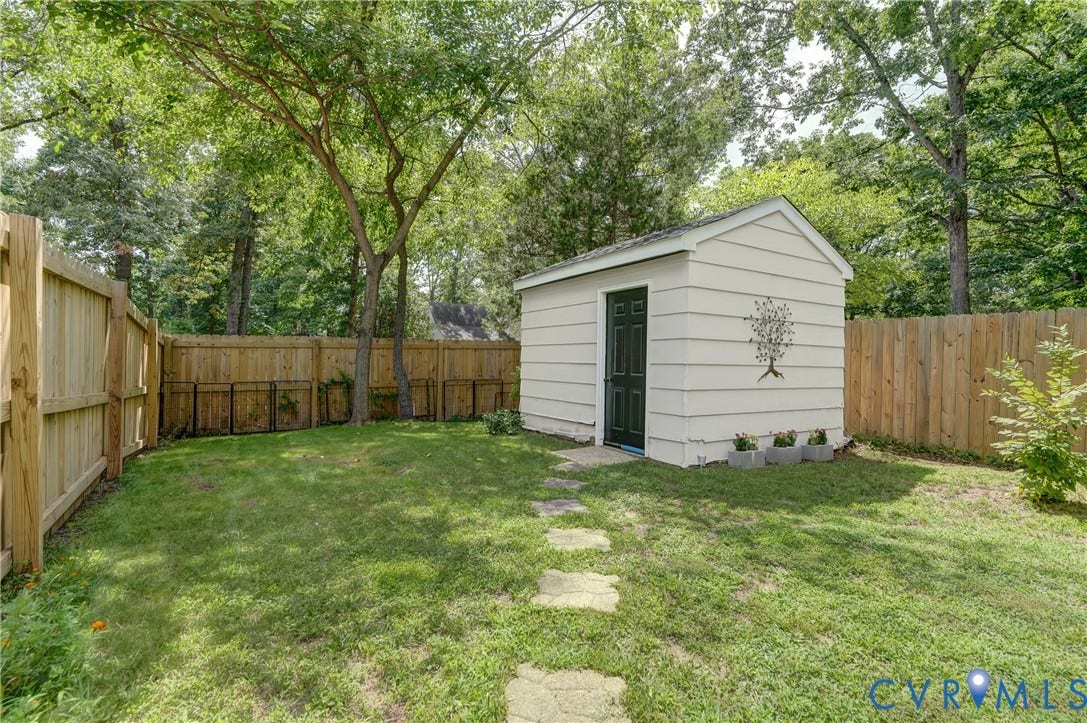 Expansive grassy yard with a wooden privacy fence, mature trees, a storage shed with a dark-paneled door, and stone pathway sections