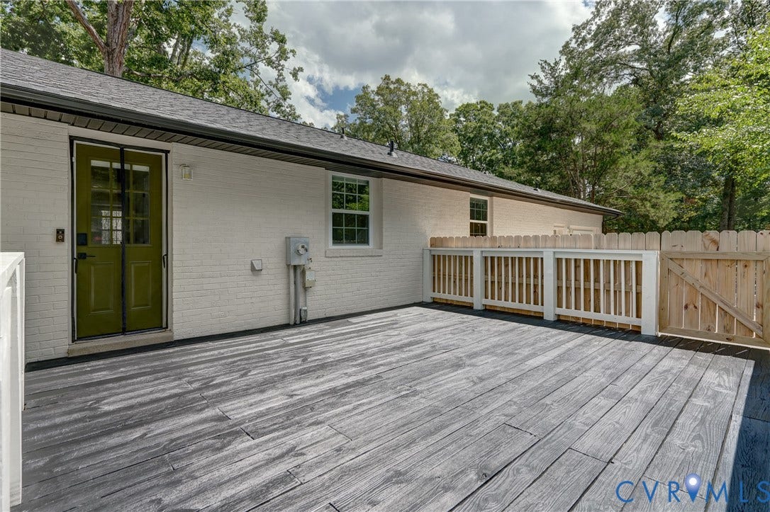 Expansive outdoor deck with wood-finish flooring, featuring a white brick exterior, a green entry door, and a privacy fence with a gate