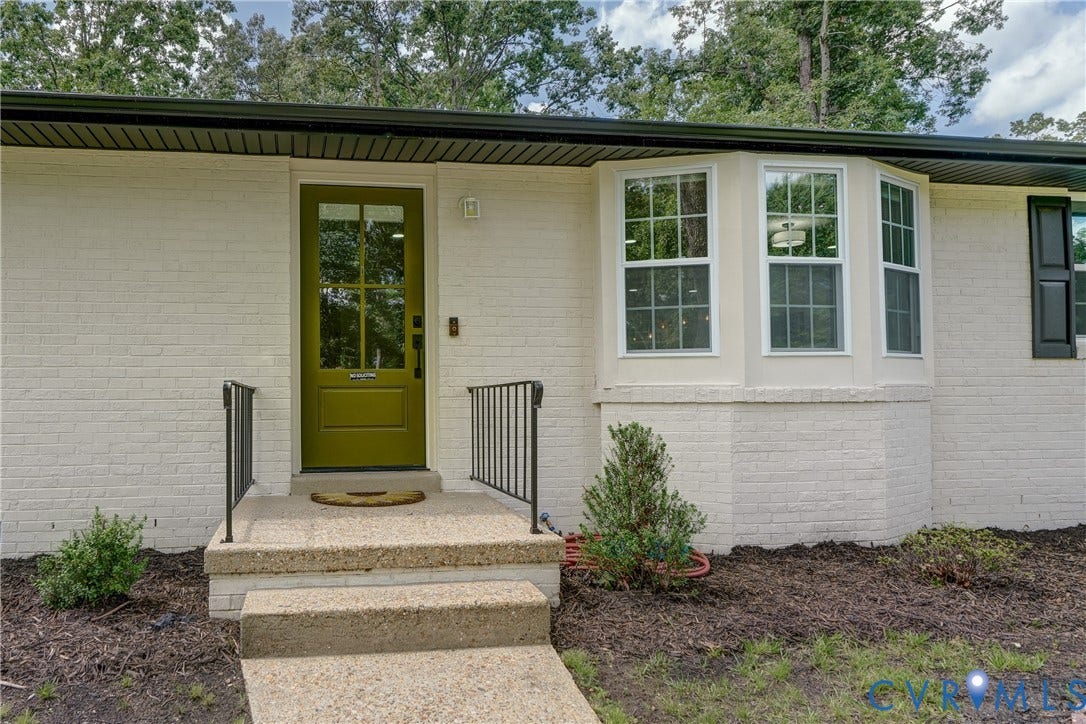 White painted brick exterior with a green paneled entry door, bay window with multiple panes, black gutters, and a black wrought iron handrail