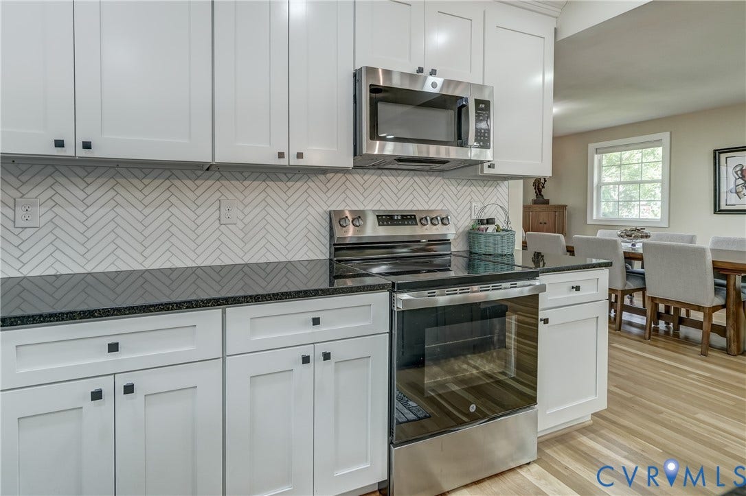 White shaker-style cabinetry with square hardware, herringbone tile backsplash, dark countertops, stainless steel appliances, and wood-finish flooring