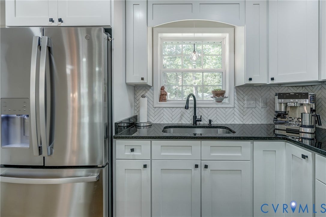 Stainless steel French door refrigerator, white shaker-style cabinetry, black countertop, black sink with a matte black faucet, and white herringbone tile backsplash