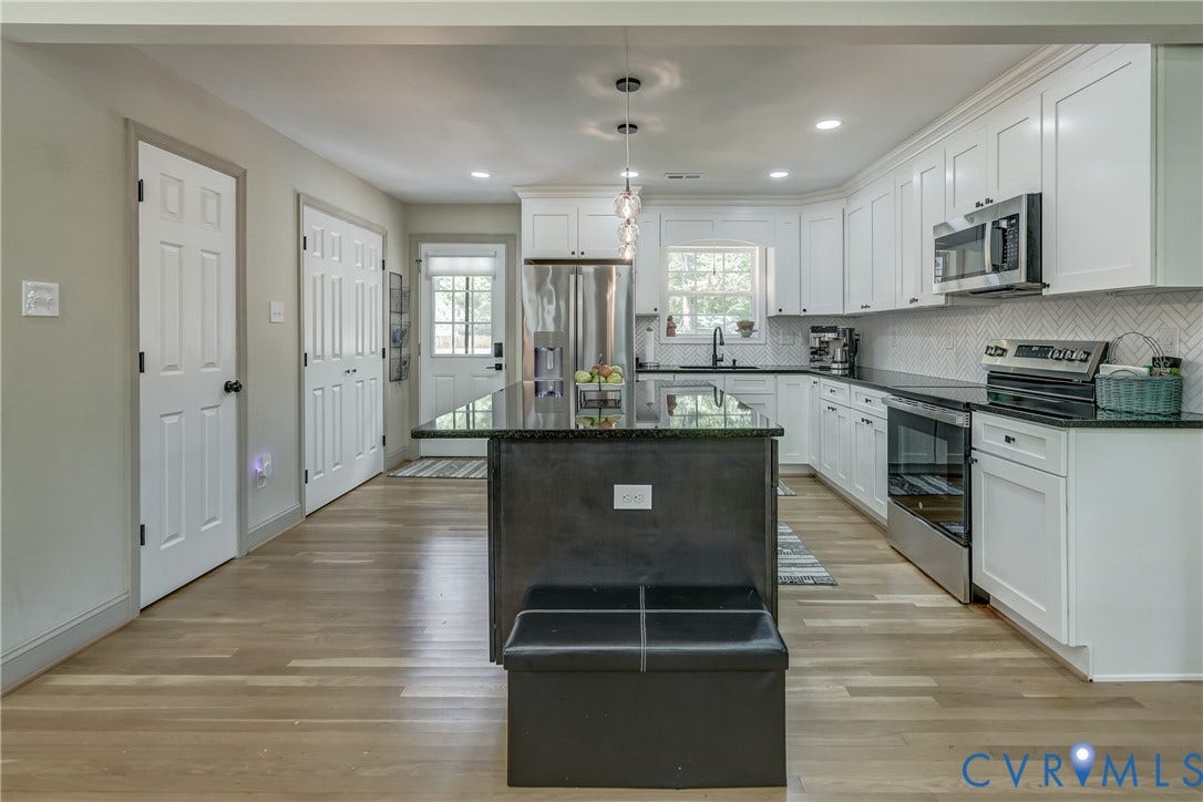 Open-concept kitchen featuring an island with a dark countertop, white shaker-style cabinetry, stainless steel appliances, a tile backsplash, and light wood-finish flooring