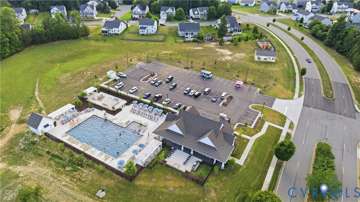 Community recreation area featuring a large swimming pool, expansive pool deck with multiple chaise lounges and umbrellas, a modern clubhouse with a covered patio, and an adjacent parking lot