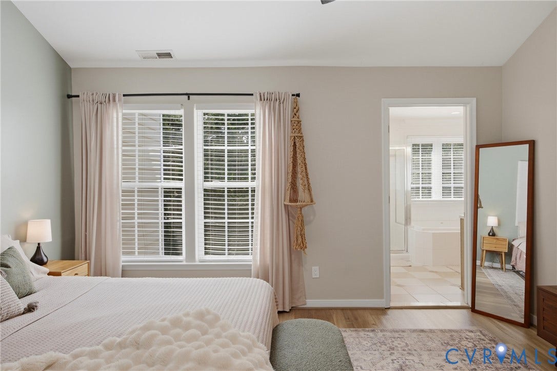 Bedroom featuring wood-finish flooring, two large windows with blinds, and an entryway to a full bathroom with tiled flooring and a built-in tub