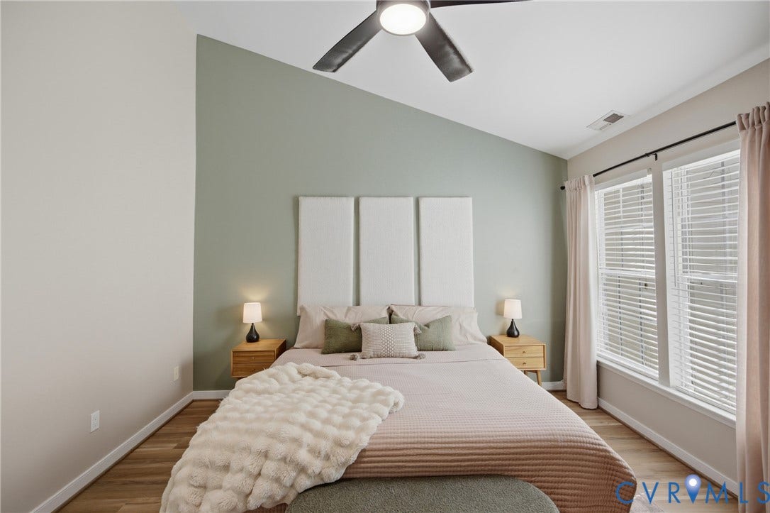 Vaulted ceiling bedroom with a modern ceiling fan, wood-finish flooring, two large windows, and a feature accent wall