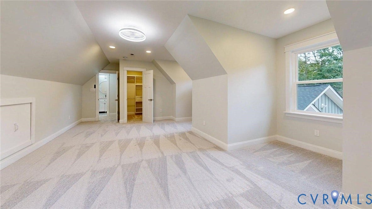 Spacious carpeted room featuring sloped ceilings, recessed lighting, a modern flush-mount light fixture, a window with exterior views, and access to a walk-in closet with built-in shelving