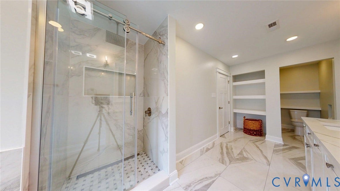 Spacious bathroom featuring a glass-enclosed shower with subway tile, built-in shelving, a dual-sink vanity with a light-toned countertop, and recessed lighting