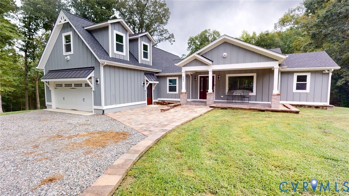 Modern farmhouse exterior with gray board and batten siding, a covered front porch featuring stone-clad columns, a paved patio, a gravel driveway, and an attached garage with a white door