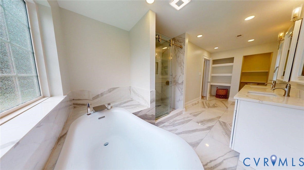 White freestanding soaking tub with deck-mounted fixtures, marble-patterned tile flooring, a glass-enclosed shower, dual vanity with undermount sinks and gold-toned fixtures, and built-in shelving