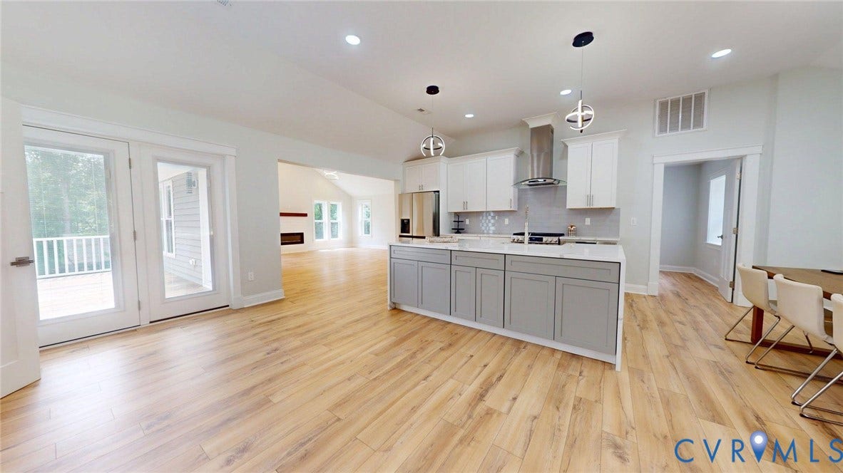 Spacious open-concept living area featuring wood-finish flooring, a modern kitchen island with seating, stainless steel appliances, recessed lighting, and double glass doors opening to an exterior deck
