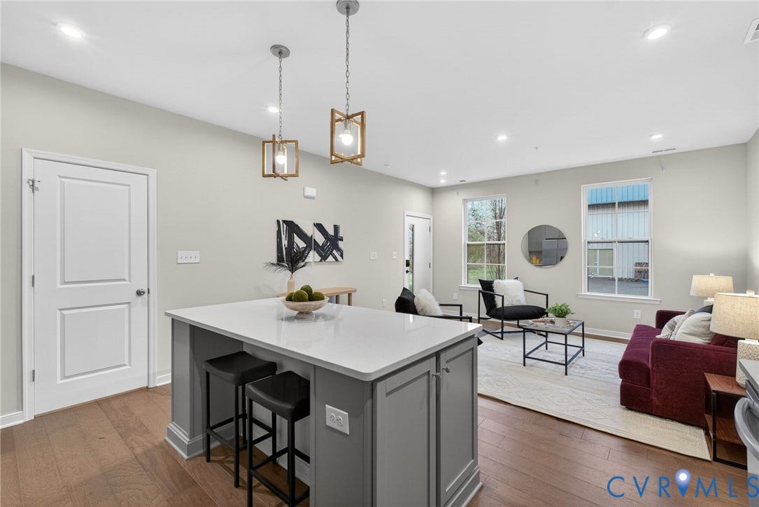 2439 Ownby Lane, #B 13, Richmond, VA 23220 Open-concept kitchen and living area featuring a gray island with waterfall countertop and seating for two, white quartz countertops, pendant lighting with brass accents, hardwood flooring throughout, and abundant natural light from multiple windows