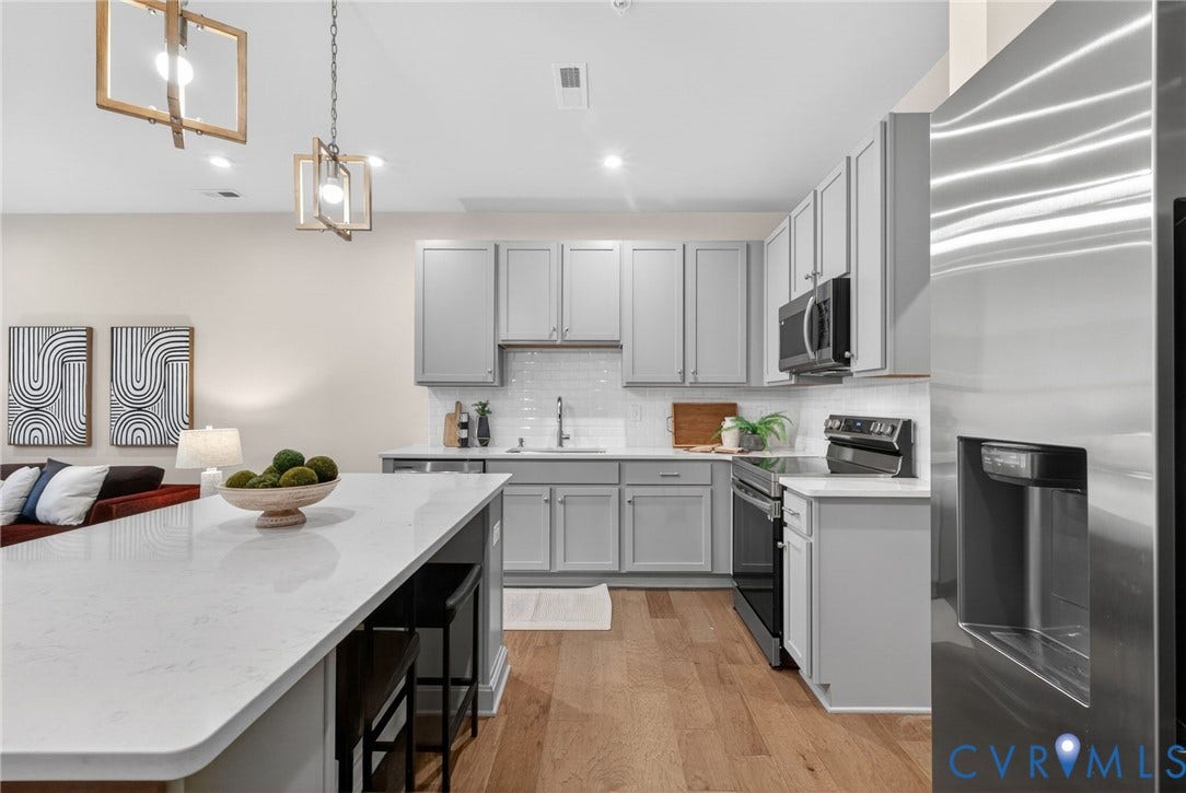 2439 Ownby Lane, #B 13, Richmond, VA 23220 Modern kitchen featuring white cabinetry, stainless steel appliances including a built-in wall oven and dishwasher, white quartz countertops, a large waterfall-edge island with seating, pendant lighting, and wood-finish flooring throughout the open layout